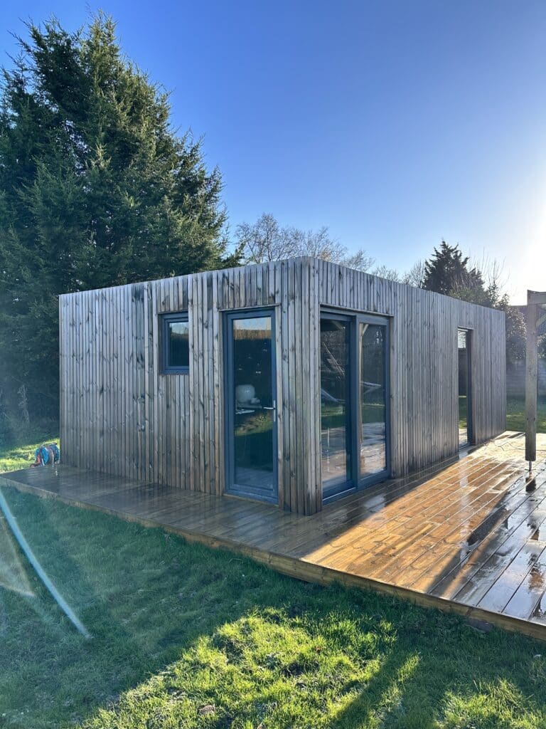 Studio de jardin à toit plat avec bardage bois et baies vitrées d'angle posé sur terrasse en bois