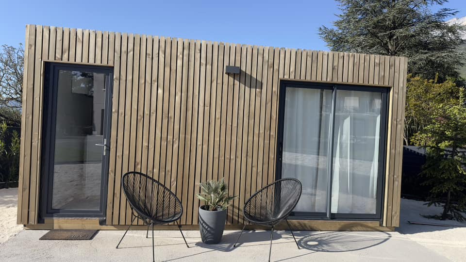 Studio de jardin en bois avec porte et baie vitrée, aménagé avec des chaises et une plante.